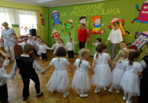 Dzieci odświętnie ubrane tańczą po kole przy piosence pt. "Paluszek".